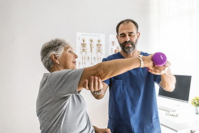 Physical therapist guiding the arm of a woman with white hair as she lifts a small hand weight in an office therapy setting.