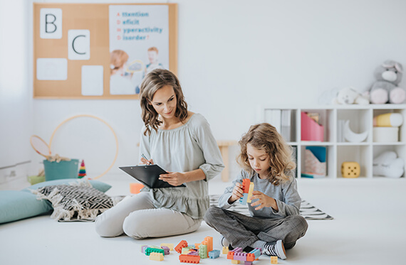 Child is sitting on the floor and playing with block. BCBA sits next to child to observe and take notes on a clipboard